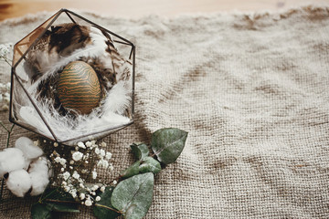Stylish easter egg in modern nest with feathers on rustic table. Natural dyed green easter egg with eucalyptus branch, spring flowers, cotton on rural background. Space for text