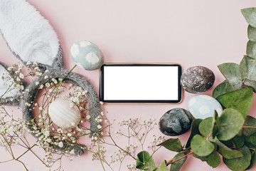 Stylish easter eggs, phone with empty screen, bunny ears, flowers border flat lay on pink wallpaper with copy space. Natural dyed marble easter eggs and grey rabbit ears. Greeting card template