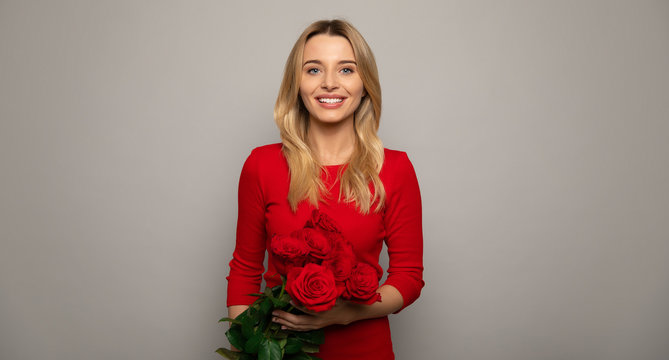 Red roses. Close up photo of a blonde woman in a red outfit, who is posing in front, holding a bouquet of red roses, smiling and looking in the camera.