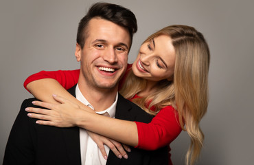 Love and beauty. Close up photo of a fantastic girl in a red dress, who is hugging her boyfriend in a suit from the back, looking at him while he is looking in the camera and smiling.