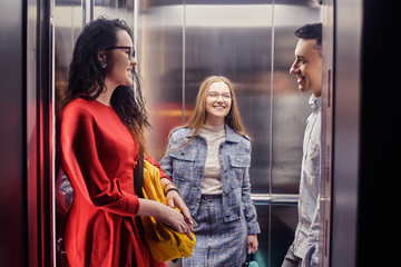The girls and the guy ride in the elevator. Students in the elevator go to study. People in the elevator. Elevator with people, communication in public places. Colleagues go to work in the elevator. - obrazy, fototapety, plakaty