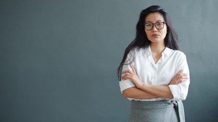 Slow motion portrait of serious Asian businesswoman standing on gray background with arms crossed looking at camera. People and lifestyle concept. - Powered by Adobe