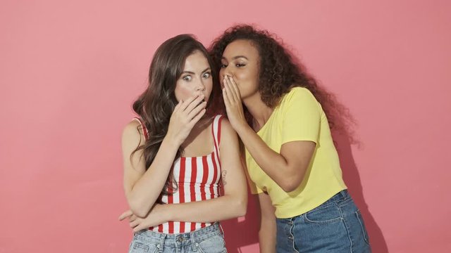 Two mystery pretty girls friends having secret and whispering among themselves while one of them having shocked after that over pink background