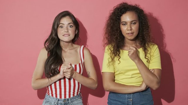 Two intrigued pretty girls friends experiencing excitement and becoming sad after that over pink background