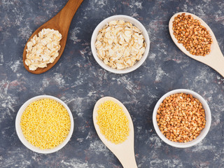 Groats buckwheat, oatmeal, millet in white cups and wooden spoons on a dark concrete background. Weight loss concept