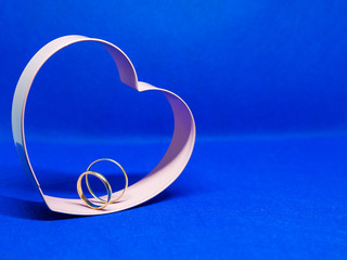 Heart-shaped cookie mold frame. In the center wedding rings. Blue background, isolated, copy space...