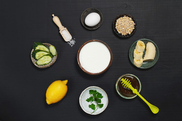Ingredients for kefir face mask: kefir in a bowl, banana, honey, parsley, lemon, cucumber, sea salt, egg, oatmeal, on a dark background in ceramic saucers.Horizontal position, top view.