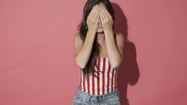 Smiling pretty brunette woman covering her face and then put her hands away and becoming happy over pink background