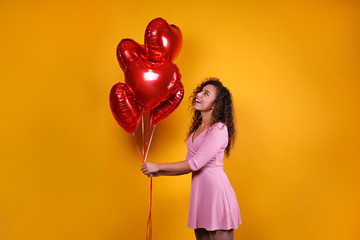 Studio portrait of young woman with dark skin and long curly hair wearing sexy dress over the festive red wall with heart shaped balloon. Close up, isolated background, copy space.