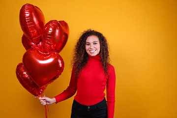 Studio portrait of young woman with dark skin and long curly hair wearing sexy dress over the festive red wall with heart shaped balloon. Close up, isolated background, copy space.