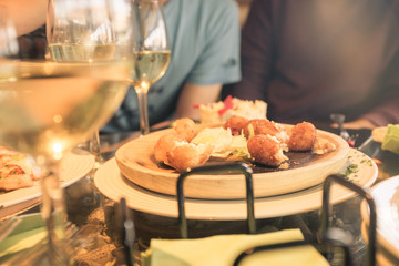 Plate of food and glasses of wine during a meal of friends. Several croquettes placed on a wooden plate with glasses of wine on glass table, with two friends in unfocused background