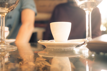 White cup of coffee on a glass table with glasses of wine and two Caucasian young boys on unfocused background with soft sunlight entering from the side