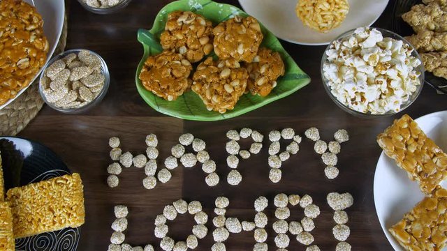 Zoom out shot of 'Happy Lohri' text written with rewri on a wooden table in India. Top view shot of Lohri items like gajak  rewri  peanuts chikki  bhujia chikki  murmura ladoo  and popcorns consume...