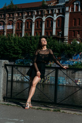 beautiful brunette stands on the bridge, near the river,in the city. a girl in a black dress puts a face to the sun and the wind that blows on her