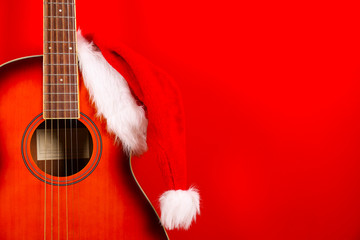 Santa Clauses hat hanging on vintage style travel size acoustic guitar with rosewood neck and no pickguard over festive red wall background. Close up, copy space for text, top view.