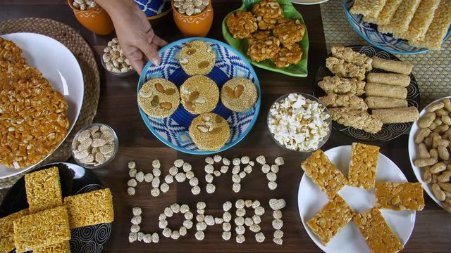 Woman hands decorating a wooden table with Lohri items consumed in winters. Top view shot of Happy Lohri text surrounded by til patti  peanuts chikki  rewri  popcorns  gajak rolls  groundnuts  etc ...