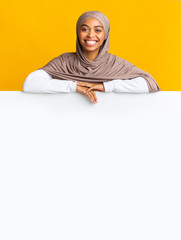 Smiling black muslim girl in headscarf leaning on white advertisement board