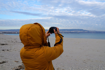 woman in a yellow jacket photographs an empty beach and the sea in winter