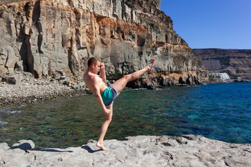 Red hair man training kickboxing by rocky beach in Puerto Mogan, Gran Canaria. Young sports man practicing martial arts by the sea in Canary Islands, Spain. Healthy lifestyle concept