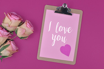 Romantic Valentine's Day flatlay with clipboard with pink paper and words 'I love you' and heart next to rose flower bouquet on magenta background