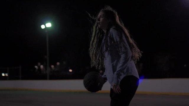 Low angle tight shot of teenage girl dribbling basketball then taking a shot at an outdoor court at night with lights.