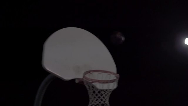 Teenage girl dribbles ball then shoots it at net at an outdoor court at night.