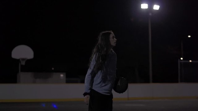 Teenage girl with long brown hair dribbles ball, throws it up and catches it before shooting at a dark outdoor court with lights.