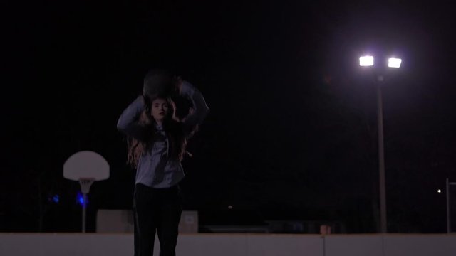 Cinematic shot of teenage girl shooting a basketball in the net before lights in a dark outdoor court.
