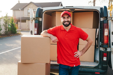 Image of smiling young delivery man standing with parcel boxes near car