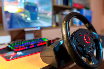 Truck driving simulator steering wheel on focus in front of a computer with glowing keyboard,...
