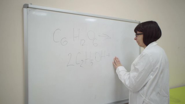 Chemist student in white lab coat trying to write simplified formula for the synthesis of ethanol from glucose, but forgot from frustration banging his head on the board.