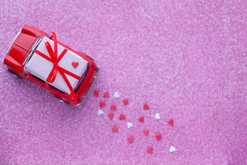 Red toy car delivering carrying on roof gift box with hearts on pink background. Valentine's day concept.