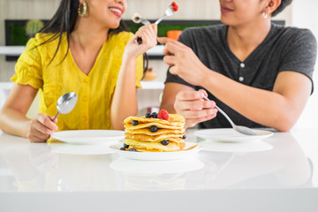 Young Asian man and woman couple feeding each other dessert with a plate of stack pancake on the table, main focus on pancake.