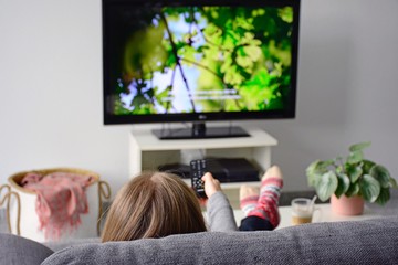 Young woman watching television with subtitles while sitting comfortably on sofa at home in living room. Nature, green, documentary, tv screen - obrazy, fototapety, plakaty