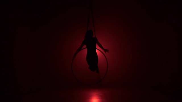 Silhouette Aerial acrobat in the ring. A young girl performs the acrobatic elements in the air ring on red background. Slow motion. 056