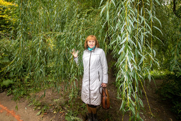 Russian woman in white raincoat cost(stand)s under branch of the osier