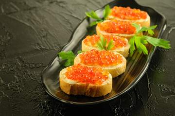Red fresh caviar applied on the slices of white loaf on the black dish and on the black background.