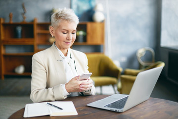 Modern lifestyle, communication and electronic gadgets concept. Busy attractive elderly female entrepreneur with pixie blonde hair working in office with laptop and notes on desk, using smart phone