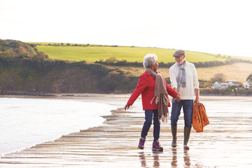 Senior Couple Hold Hands As They Walk Along Shoreline On Winter Beach Vacation