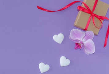 Gift box, orchid flower and white hearts on a violet background. Valentine's day and romantic concept. Flat lay. Copy space.