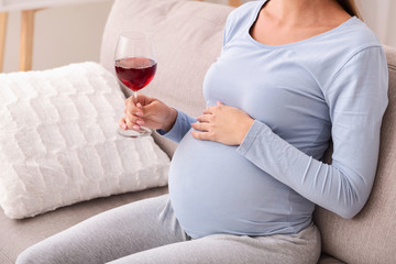 Unrecognizable mother-to-be holding glass of wine sitting on sofa, cropped