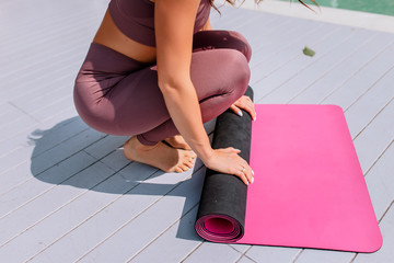Yoga mat and meditation. Young woman rolls a pink mat after yoga class on a white wooden floor. Girl in a lilac set. Classes on the background of the sea. The girl folds the rug.