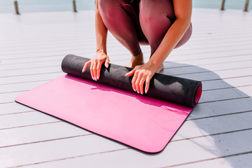 Yoga mat and meditation. Young woman rolls a pink mat after yoga class on a white wooden floor.  Girl in a lilac set. Classes on the background of the sea. The girl folds the rug. Top view.