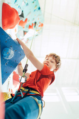 Teenager boy at indoor climbing wall hall. Boy is climbing using a top rope and climbing harness...