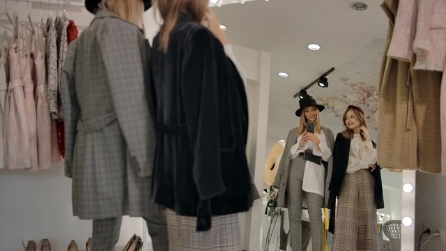 Slow motion, pan right of female friends standing in front of mirror and taking photo with smartphone while trying on stylish clothes in fashion boutique.
