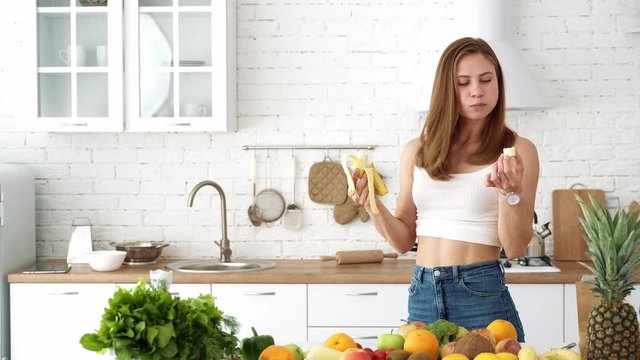 Cheerful girl eats a banana in a white kitchen with a table with vegetables and fruits.