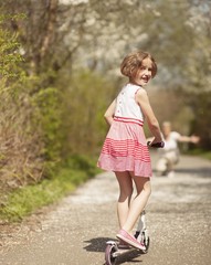 Young girl riding scooter in park away from camera to mother