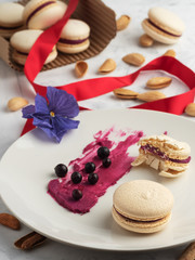 Obraz na płótnie Canvas Homemade macarons with currant filling on a gray plate. Selective focus on cookies. Almonds, berries of blackcurrant and filling for cookies on a plate. Close-up. Red ribbon for gift wrapping.