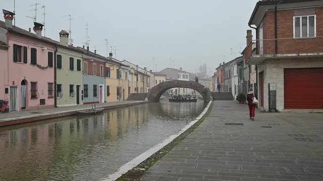 Comacchio, Italy. December 2019. Shot on a beautiful canal: houses are reflected in the water. A woman walks along the bank. In the background, a bridge surrounded by fog is crossed by people.