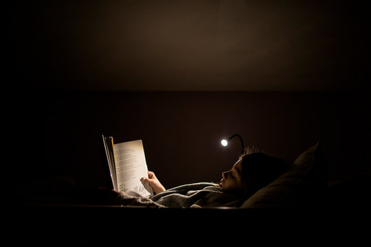 Backlit image of young girl reading by lamp light in bed at nighttime